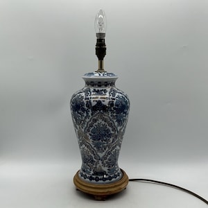 May include: A table lamp featuring a ceramic vase-shaped base decorated with a blue and white floral pattern and gold accents. The lamp sits on a circular wooden base and includes a metal socket with a clear light bulb extending from the top.