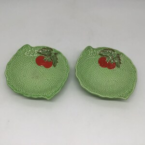 May include: Two matching ceramic plates shaped like green leaves with textured veins. Each plate features a raised, painted design of two bright red tomatoes with dark green leaves positioned near the top edge of the leaf-shaped surface.