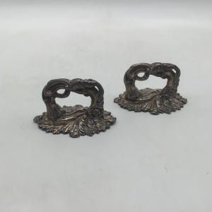 May include: A pair of antique silver-coloured drawer pulls. Each pull has an open, decorative handle with intricate detailing, set on a base with a leaf design. The pulls are shown against a plain, light background.