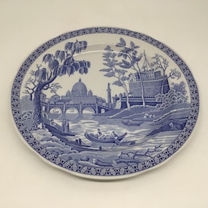 May include: Oval ceramic plate with a blue and white scenic design. The plate features a detailed illustration of a European cityscape with a river, bridge, and buildings. The rim has a decorative border.