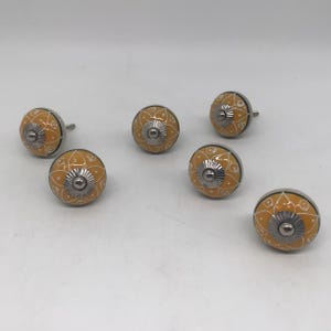 May include: Six yellow and white ceramic cabinet knobs with a floral design. Each knob has a silver metal base and stem. The knobs are arranged on a white surface.