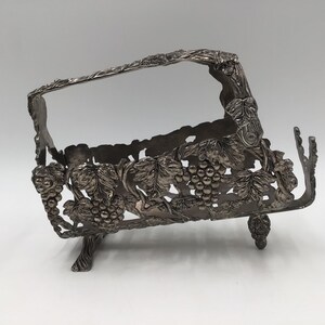 May include: A silver-toned metal basket featuring an intricate relief design of grape clusters and vine leaves. The basket has an arched handle, an openwork body, and stands on four small feet shaped like bunches of grapes, with a metallic finish.
