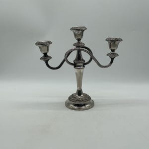 May include: A silver-toned metal candelabra featuring three candle holders arranged in a symmetrical design. The central stem is flanked by two curved arms, with decorative embossed detailing around the base and the rims of each candle cup.