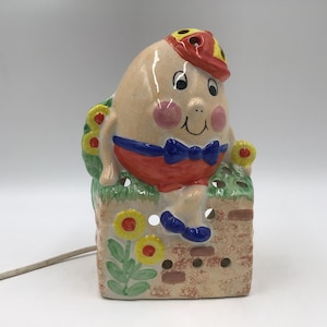 May include: A ceramic Humpty Dumpty night light. The egg-shaped character wears a red hat, shorts, and a blue bow tie. The base is a brick wall with painted flowers and greenery. The night light has a power cord.