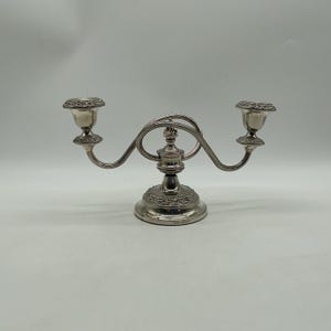 May include: A silver-toned metal candelabra featuring two candle holders connected by curved arms to a central pedestal base. The base and the rims of the candle cups are decorated with a detailed embossed floral pattern and a smooth finish.