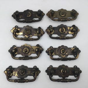 May include: Eight antique brass drawer pulls. Each pull has a decorative backplate with a central circular design and two small knobs. The pulls have a curved handle and show signs of age and wear, with a tarnished finish.