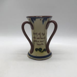 May include: A cream-coloured ceramic vase with two curved handles, featuring brown and blue accents. The vase has the text "Half of the ills we hoard within our hearts" in brown lettering. A decorative green swirl is near the base.