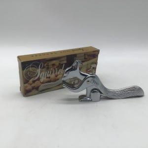 May include: A shiny chrome-plated metal nutcracker shaped like a squirrel, featuring a hinged jaw mechanism and a textured tail. It is positioned next to its original rectangular cardboard box, which displays the text Squirrel Nut Cracker.