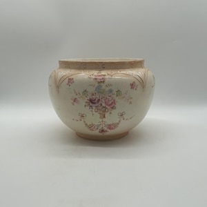 May include: A cream-coloured ceramic bowl featuring a rounded silhouette with a wide opening. The exterior is decorated with a central floral bouquet in shades of pink, purple, and blue, accented by delicate ribbons and embossed scalloped edges.