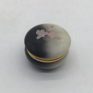 May include: A round, lidded trinket box featuring a smooth, gradient finish that transitions from deep black to off-white. The lid is decorated with a delicate, hand-painted floral motif in soft pink and white tones, accented by a thin gold-tone metal rim.