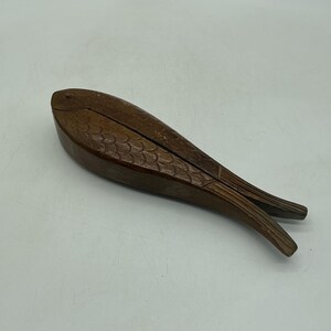 May include: A dark brown wooden nutcracker shaped like a fish. The surface features carved scale patterns along the body and a defined tail section. The hinged mechanism allows the fish to open and close, with a visible seam running along the top.