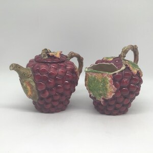May include: A ceramic teapot and creamer set. The teapot and creamer are shaped like bunches of grapes, with a deep red colour and textured surface. The handles and spout are designed to resemble branches.