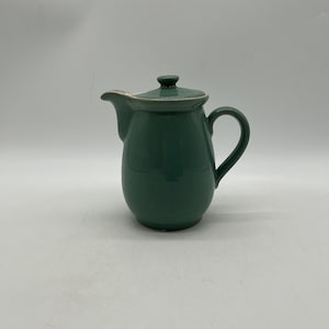 May include: A sage green ceramic jug with a matching lid, a curved handle, and a wide pouring spout. The jug features a smooth, glossy finish with a subtle light-coloured rim detail along the lid and the top edge of the vessel body.