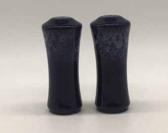 May include: A pair of navy blue salt and pepper shakers. The shakers have a flared shape with a speckled, lighter blue design near the top. The shakers are ceramic and have a glossy finish.