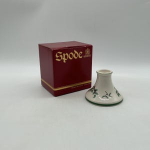 May include: A white ceramic candle holder with a flared base and a holly leaf and berry pattern, featuring a green rim. Beside it sits a square, deep red cardboard box with the gold text Spode and Stoke-on-Trent England printed on it.