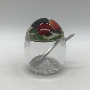 May include: A clear glass sugar bowl with a decorative lid featuring a strawberry design. The lid is painted with red strawberries, green leaves, and a black background. A small silver spoon rests inside the bowl.