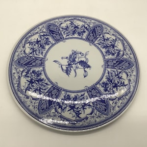 May include: A round ceramic plate featuring a white base with a blue transferware pattern. The design includes a central holly sprig surrounded by a border of alternating panels containing holly leaves, berries, wheat stalks, and pine cones.