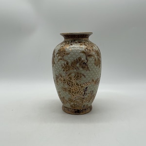 May include: A ceramic vase featuring a light green base with a repeating white geometric pattern. The surface is decorated with intricate gold floral designs, including large blossoms and leafy vines, with gold bands at the rim and base.