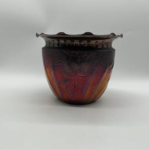 May include: A dark, iridescent metal planter with a ruffled, scalloped rim. The exterior features a hammered texture and an embossed floral pattern of tulips with long, slender leaves. The finish transitions from deep purple to copper and bronze.