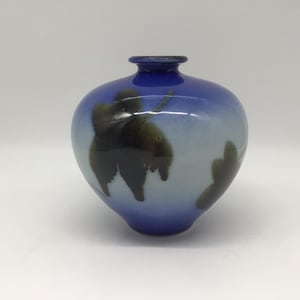 May include: A small, rounded glass vase featuring a smooth, glossy finish. The body transitions from a deep, saturated blue at the top and bottom to a lighter, pale blue in the centre, decorated with dark, abstract, leaf-like shapes in black.