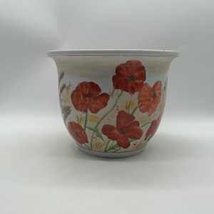 May include: A white ceramic plant pot featuring a hand-painted botanical design of vibrant red poppy flowers with green stems and foliage. The pot has a wide, flared rim and a rounded body, showcasing a repeating floral pattern against a cream background.