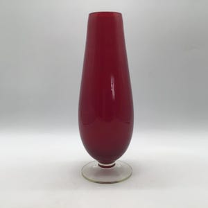 May include: A tall, smooth, deep red glass vase with a tapered, teardrop-shaped body that narrows towards the top opening. The vessel is supported by a clear, circular glass pedestal base, providing a stable foundation for the vibrant red structure.