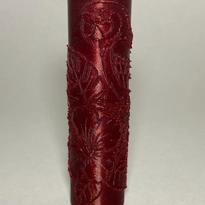 May include: A cylindrical red paint roller with a raised, embossed pattern featuring a tropical design of a parrot, hibiscus flowers, and large monstera leaves. The surface has a metallic, shimmering finish and a hollow center opening.