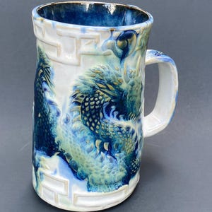 May include: A ceramic mug featuring a raised, sculpted dragon design in shades of deep blue, teal, and white. The exterior surface displays a textured, relief-style dragon motif wrapping around the body, complemented by a geometric border pattern.