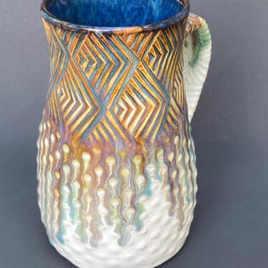 May include: A ceramic pitcher featuring a deep blue interior and a textured exterior. The upper section displays carved geometric diamond patterns in brown and blue, while the lower portion transitions into white with raised, drip-like textures.