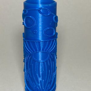 May include: A cylindrical blue 3D printed roller featuring a raised relief pattern of a gnome with a long beard and a hat decorated with circular shapes. The object has a hollow center and a smooth top edge, showcasing a monochromatic blue finish.