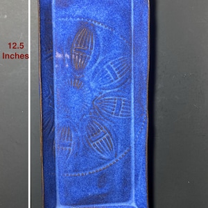May include: A rectangular ceramic tray in a vibrant blue glaze, measuring 12.5 inches long and 5 inches wide. The tray features an embossed floral pattern and a dark brown rim. The design is symmetrical, with the floral pattern centered on the tray.