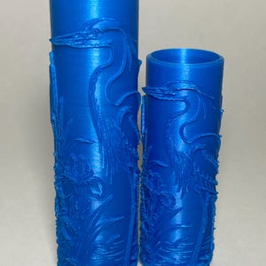 May include: Two cylindrical blue vases of different heights featuring a raised relief design of a long-necked bird standing among tall grasses and flowers. The surface has a smooth, matte finish with intricate textures depicting the wildlife scene.