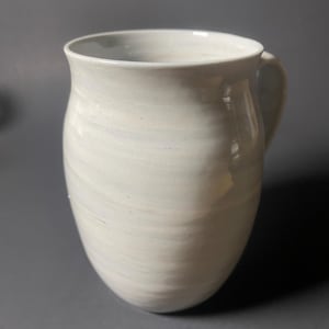 May include: A handmade, off-white ceramic pitcher with a rounded body and a curved handle. The pitcher has a slightly flared rim and a smooth, glossy finish. The surface shows subtle, swirling patterns, adding to its unique, artisanal appeal.