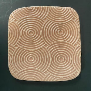 May include: A square, ceramic coaster with a repeating concentric circle pattern. The coaster is a light peach color with a glossy finish. The raised circles create a textured surface, adding visual interest to the design.