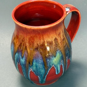 May include: A ceramic mug with a rounded body and a curved handle, featuring a glossy glaze that transitions from a deep red at the rim and base to a blend of golden brown and vibrant turquoise blue drips running down the center.