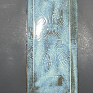 May include: Rectangular ceramic tray with a blue glaze and a floral design. The tray has a raised edge and a brown rim. The floral design is white and is in the center of the tray. The tray is approximately 10 inches long and 5 inches wide.