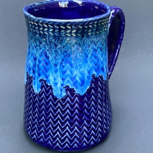 May include: A ceramic mug featuring a deep blue base with a textured chevron pattern. The upper section transitions into a lighter, vibrant blue glaze with a braided rim detail. The mug includes a sturdy handle on the right side and a smooth interior.
