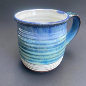 May include: A handmade ceramic mug with a blue and white glaze. The mug features horizontal stripes and a curved handle. The interior is white, and the rim is dark blue. The mug is approximately 4 inches tall.