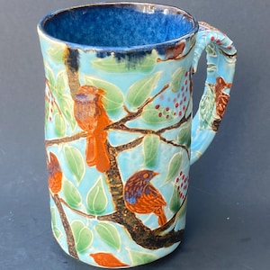 May include: A ceramic mug with a light blue background featuring a design of birds, leaves, and berries. The birds are orange and brown, and the leaves are green. The handle is also decorated with the same design.