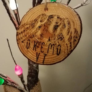 May include: A wooden Christmas ornament with the words "Okemo VT" burned into the surface. The ornament is hanging from a branch with white faux snow and colorful Christmas lights.