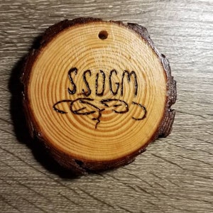 Peut inclure: Une décoration ronde en bois avec le texte "SSOGM" brûlé à la surface. La décoration a un motif de grain de bois naturel et un bord extérieur brun foncé.