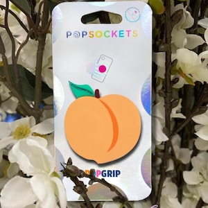 May include: A peach-shaped PopSocket phone grip on a white and iridescent backing. The PopSocket has a light orange color with a darker orange stripe. The packaging has the PopSockets and PopGrip logos. The background is white flowers.