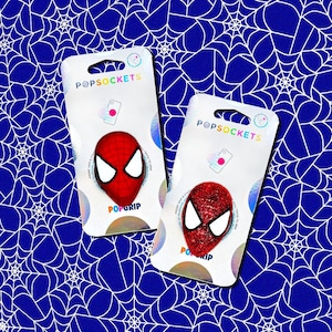 May include: Two PopSockets phone grips featuring a Spider-Man mask design. The grips are red with white eye details and are displayed on a white card with the PopSockets logo. The background is a blue spiderweb pattern.