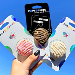May include: Three PopSockets phone grips and stands are displayed against a bright blue sky. One is a light pink, one is off-white, and the third is brown. The packaging reads "Snap better photos" and "Text with one hand."