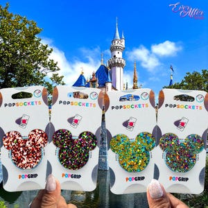 May include: Four PopSockets PopGrip phone accessories with glitter Mickey Mouse head designs. The PopSockets are red, green, gold, and iridescent. The background features a castle under a blue sky.