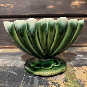 May include: A vintage green ceramic bowl with a scalloped rim and a pedestal base. The bowl is a deep green color with lighter green highlights. The base is round with a floral design and the letters "USA" are visible.