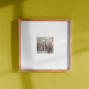Puede incluir: Una pintura de acuarela enmarcada que representa a un grupo de personas haciendo cola, posiblemente en un evento. La obra de arte está enmarcada en un marco de madera marrón claro, montado en una pared amarilla. La pintura presenta una barrera de cuerda roja.