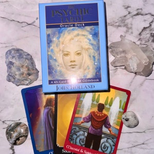 Könnte beinhalten: Ein hellblaues Tarotkarten-Set mit dem Titel "The Psychic Tarot Oracle Deck" von John Holland, mit mehreren Tarotkarten, Kristallen und Steinen. Die Karten zeigen Bilder von Menschen und Szenen.