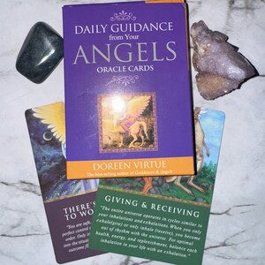 Könnte beinhalten: Eine lila Schachtel mit "DAILY GUIDANCE from Your ANGELS ORACLE CARDS" von Doreen Virtue, mit zwei Kristallen und zwei Orakelkarten. Eine Karte lautet "GIVING & RECEIVING". Die andere Karte lautet "THERE'S TO WO".