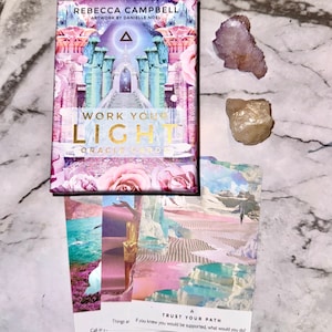 May include: A deck of "Work Your Light Oracle Cards" with a floral and architectural design, and two crystals. The card deck features the text "Work Your Light Oracle Card" in gold lettering. Several cards are fanned out below the deck, with the words "Trust Your Path" visible.